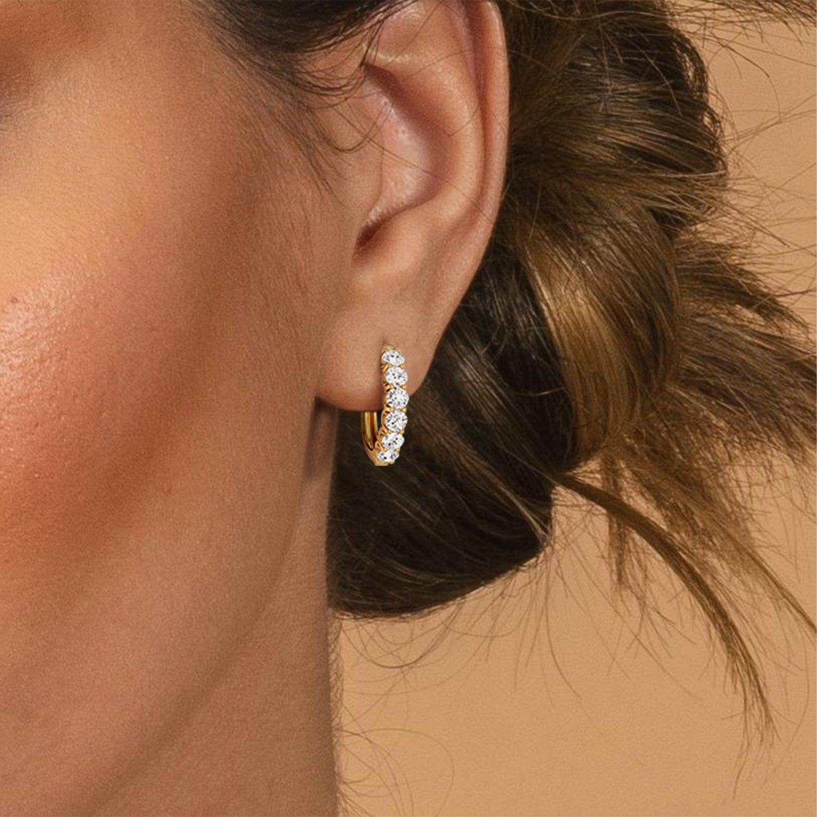 Close-up of an ear wearing a gold hoop earring with clear stones against a neutral background